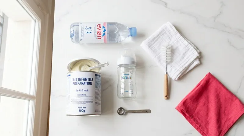 Matériel pour préparer un biberon : bouteille d'eau pour nourrissons, boîte de lait infantile, mesurette, biberon en verre et tétine sur un plan de travail propre