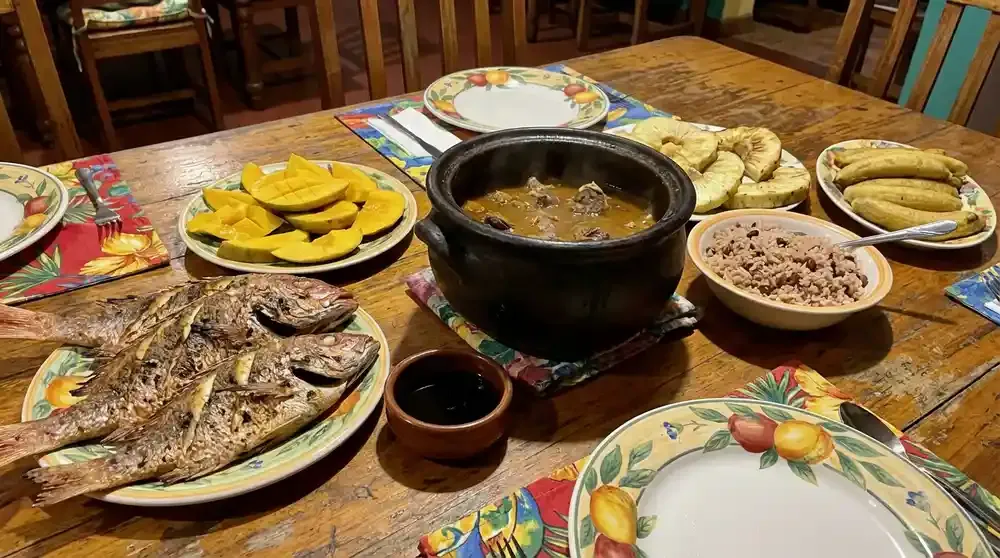 Traditional Nevisian cuisine spread on rustic wooden table featuring goat water stew in clay pot, fresh grilled fish, tropical fruits including mangoe
